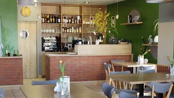 A restaurant interior with wooden tables, blue chairs, a bar area with shelves of bottles, and a green accent wall.