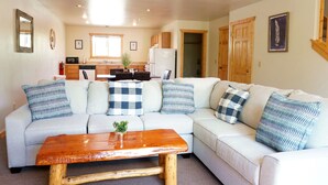 A living room with a light-colored sofa, a wooden coffee table, and a kitchen in the background. - The Aspen (West Yellowstone)