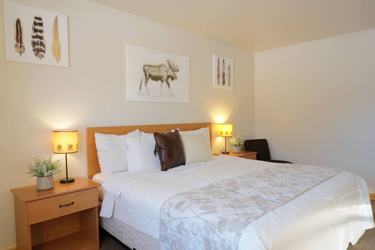 A neatly made bed with a floral bedspread, two bedside lamps, a nightstand with a plant, and wall art depicting feathers and a moose.