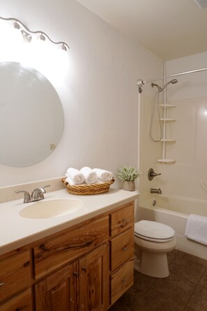 A bathroom with a wooden vanity, a round mirror, a sink, a toilet, and a shower. - The Aspen (West Yellowstone)