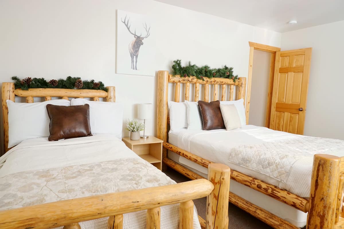 A room with two wooden beds, each with a headboard, white bedding, and a brown pillow. There is a small bedside table with a lamp and a plant. A wooden door is visible on the right.