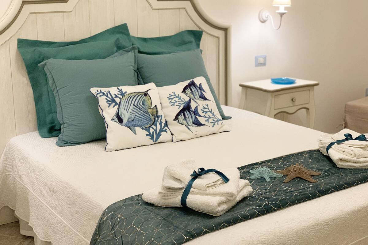 A neatly made bed with teal pillows and a decorative pillow with a fish and coral design. There is a small bedside table with a lamp and a folded towel on it.