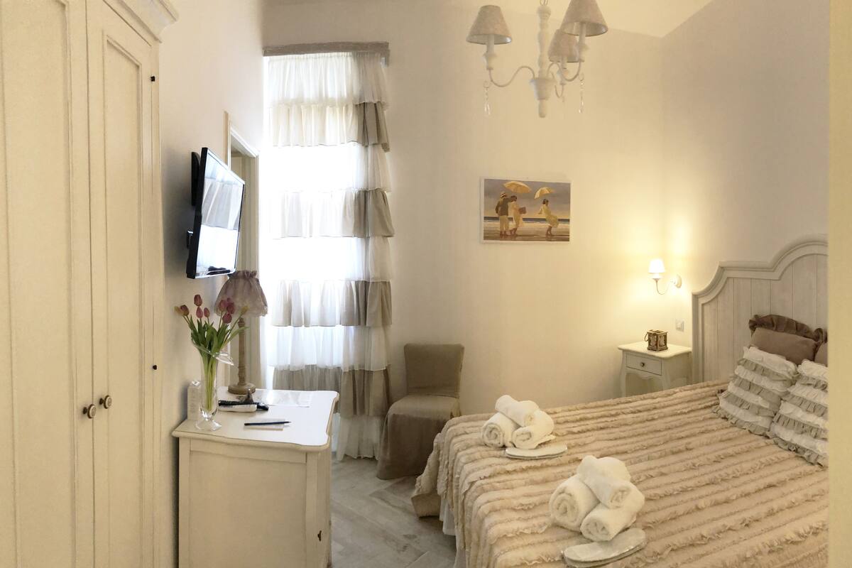 A hotel room with a bed, a desk with a vase of flowers, a chair, a painting on the wall, and a nightstand with a lamp.