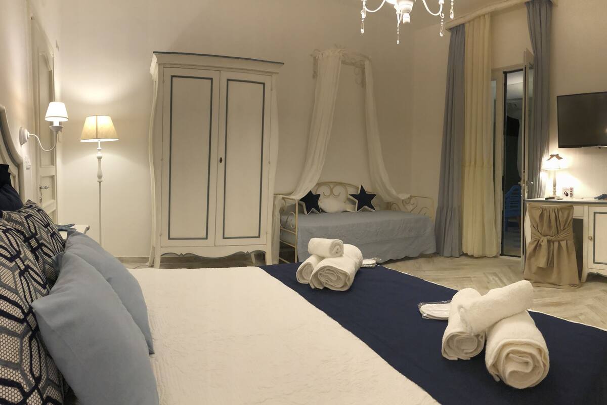 A hotel room with a bed, a wardrobe, a desk, a television, and a bathroom visible through a glass door.