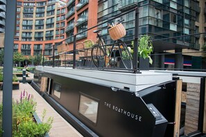 A modern boat with "The Boathouse" written on its side, docked in an urban setting with a building in the background.