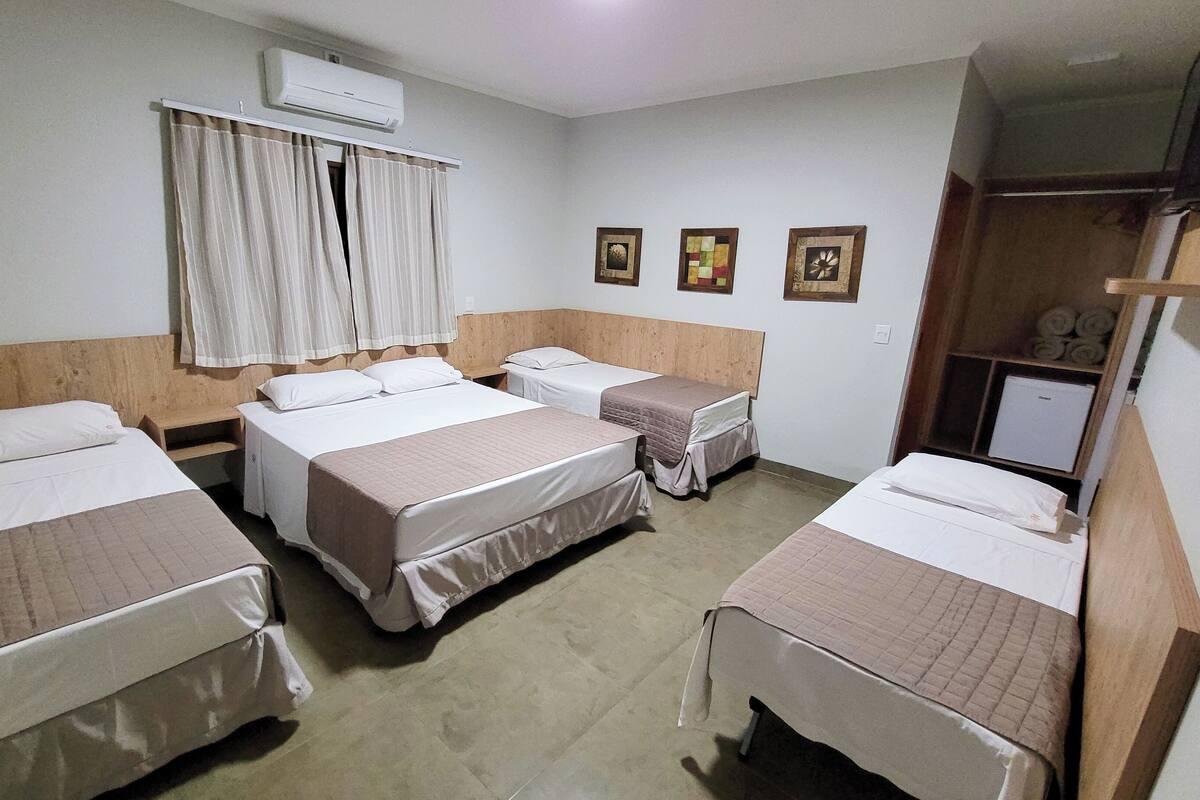 A hotel room with four single beds, a wooden headboard, an air conditioning unit, a window with curtains, and three framed pictures on the wall.