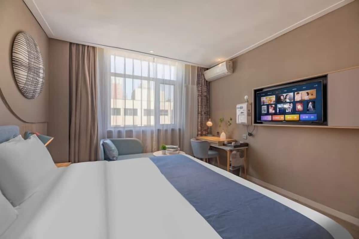 A hotel room with a large bed, a desk with a chair, a TV on the wall, and a window with curtains.