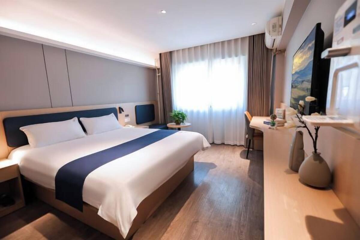 A hotel room with a bed, bedside table, desk, chair, and a large window with curtains.