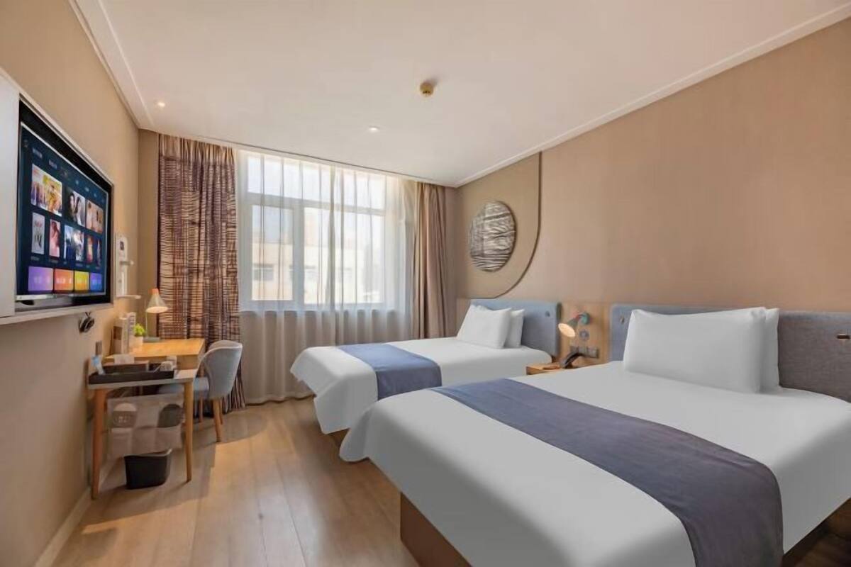 A hotel room with two beds, a desk with a chair, a TV mounted on the wall, and a large window with curtains.