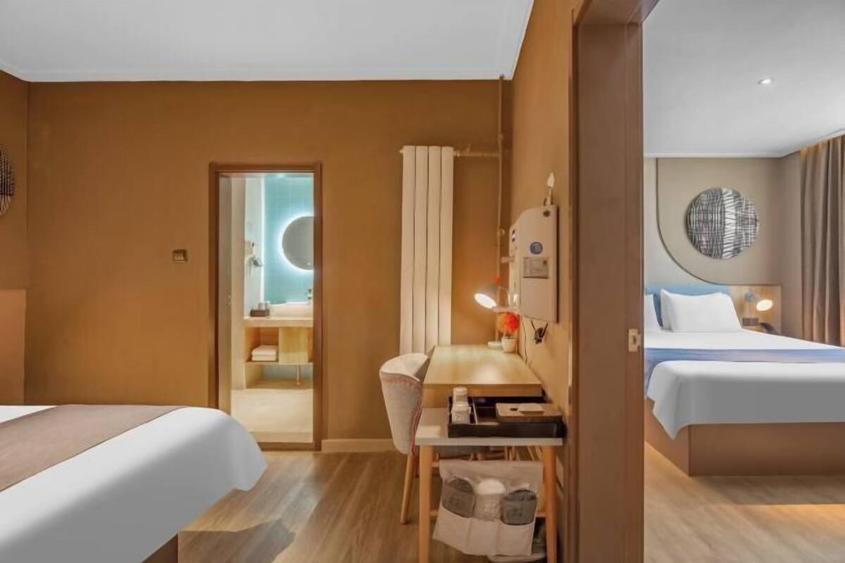 A hotel room with a bed, a desk, a chair, a mirror, a radiator, and a wall with a circular artwork.