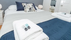 A neatly made bed with a blue and white blanket, a pillow with a geometric pattern, and a folded white towel with a bottle on it. - PML Apartments Victoria (London)