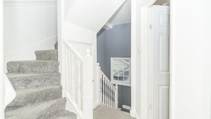 A staircase with white railings and a gray carpet leading to a room with a gray wall and a window. - PML Apartments Victoria (London)