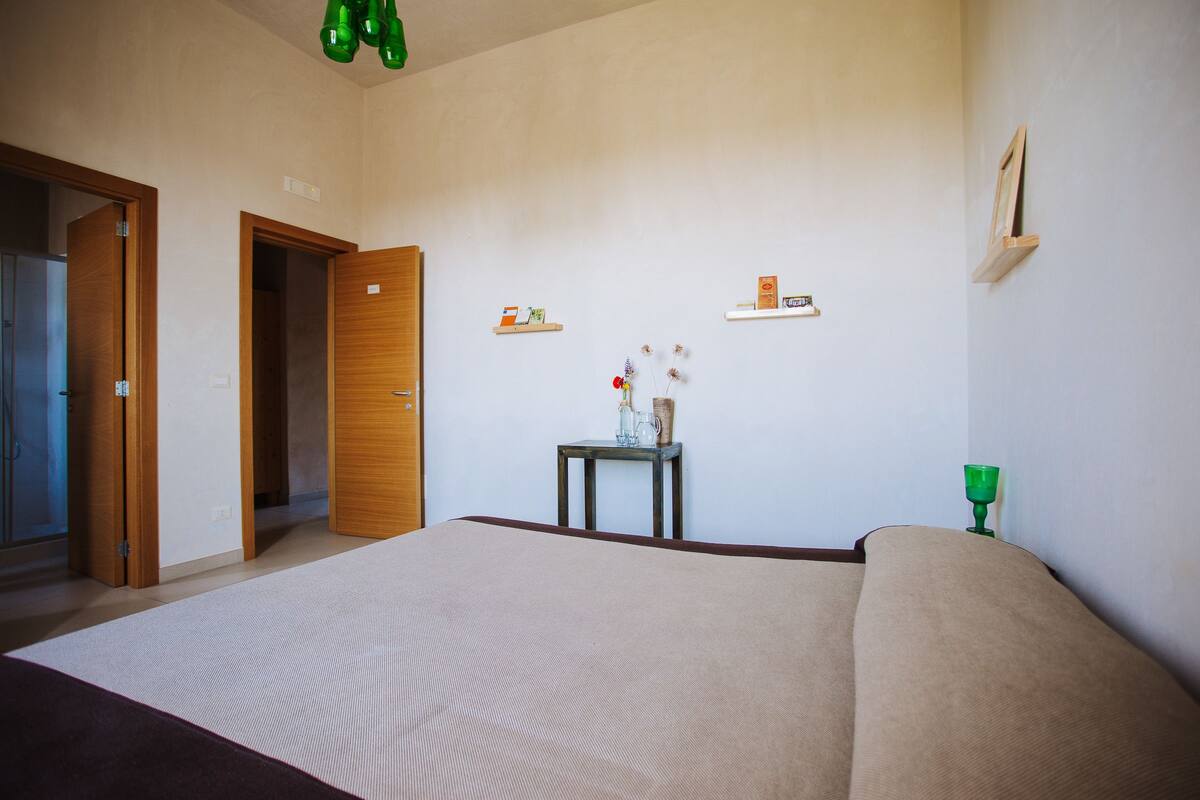 Ein Schlafzimmer mit einem großen Bett, Holztüren, einem kleinen Tisch mit einer Vase und Regalen mit Gegenständen an der Wand.