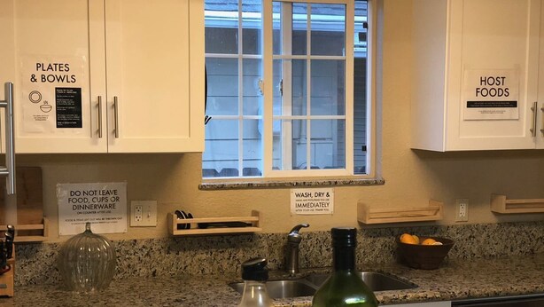 A kitchen with a granite countertop, a window, and signs indicating 'PLATES & BOWLS' and 'HOST FOODS' storage areas. - WHOLE Family - WholeStyl - Hostel (Sacramento)