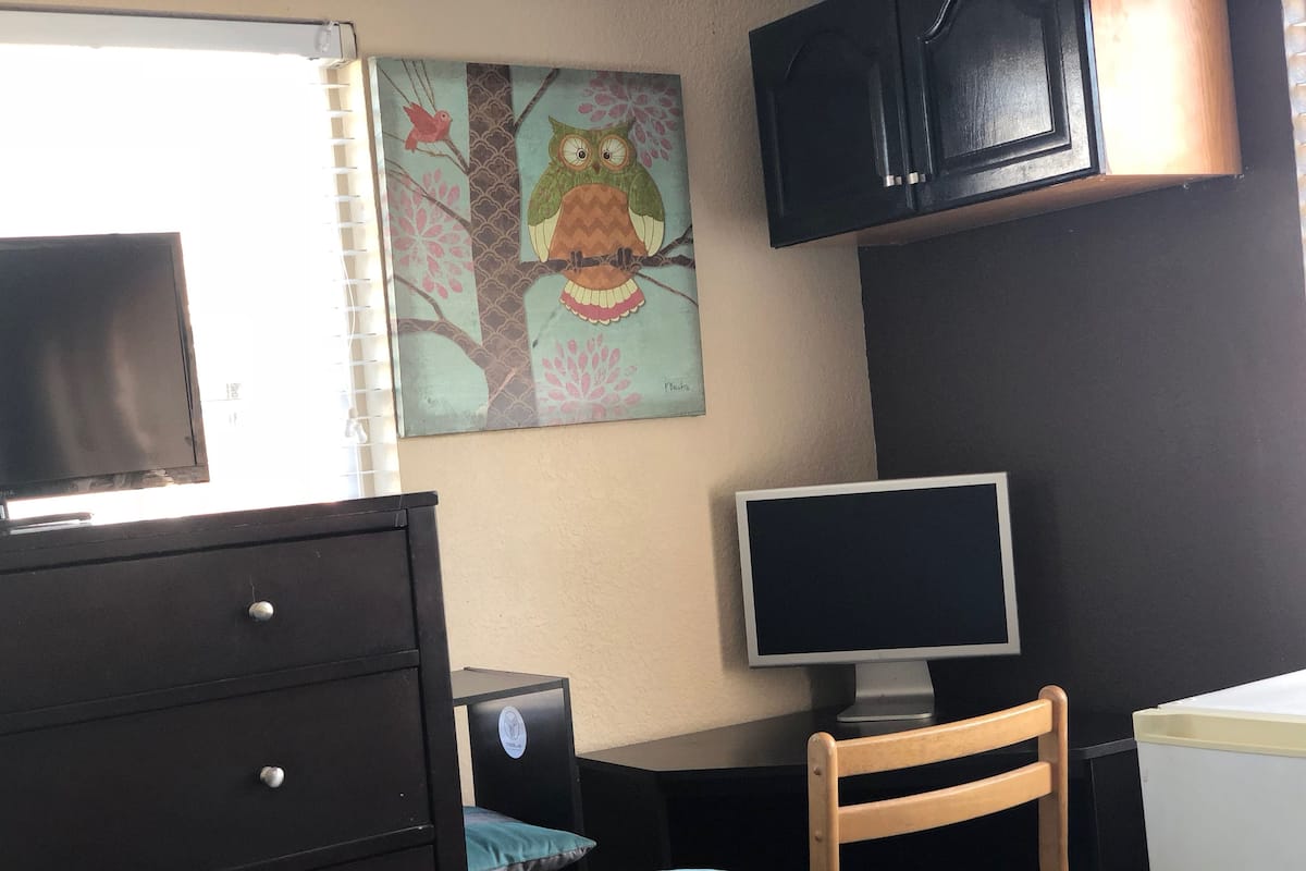 Ein Schlafzimmer mit einem Bett, einem Schminktisch, einem Schreibtisch mit Computer, einem Stuhl und einem an der Wand montierten Fernseher.