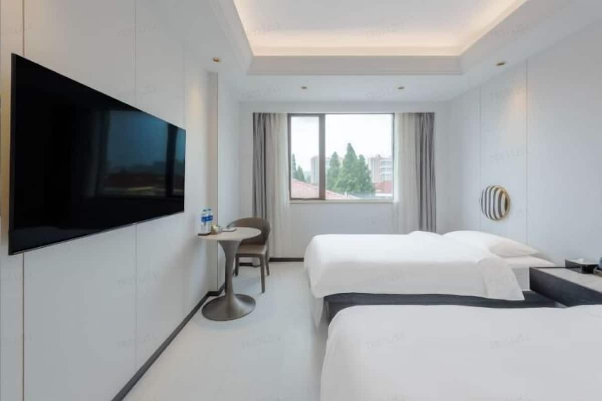 A hotel room with two beds, a TV mounted on the wall, a small table, and a window with curtains.