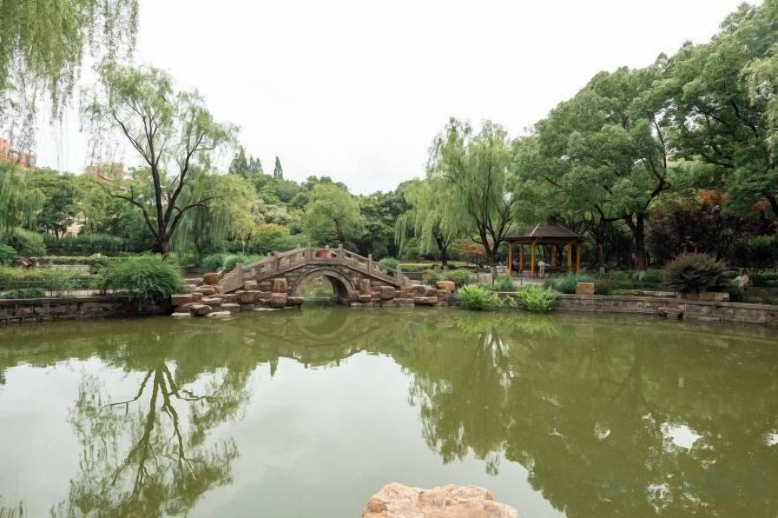 Un parque tranquilo con un puente de piedra sobre un estanque, rodeado de abundante vegetación y un pabellón.