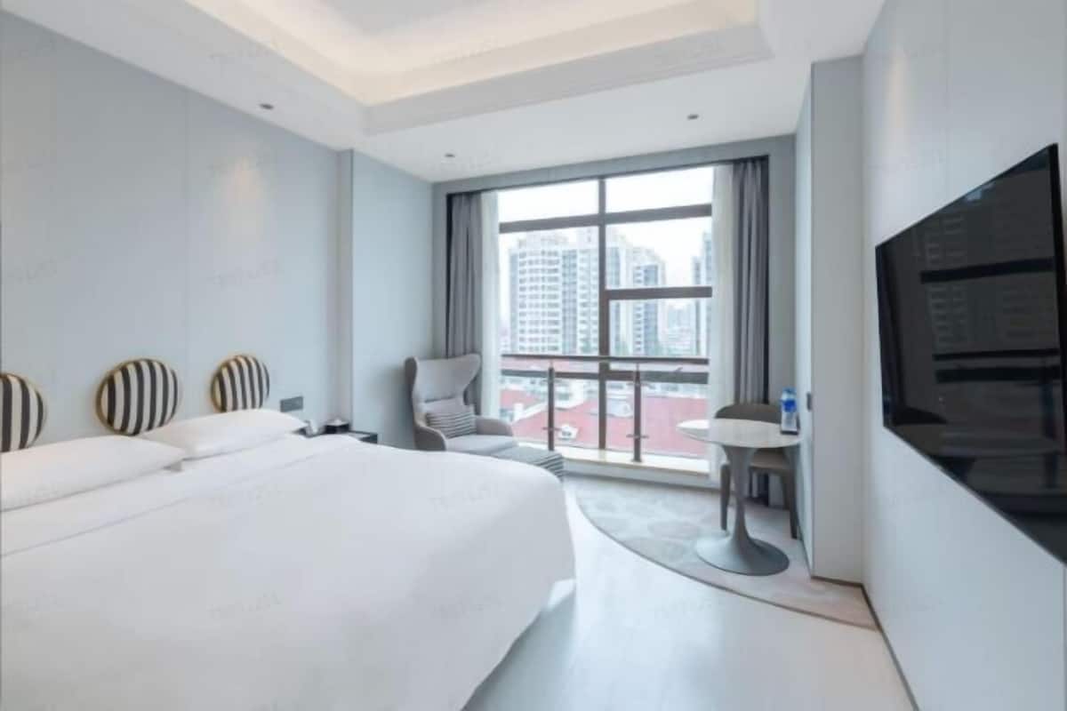 A modern hotel room with a large bed, a TV, a small table, and a view of the city through large windows.