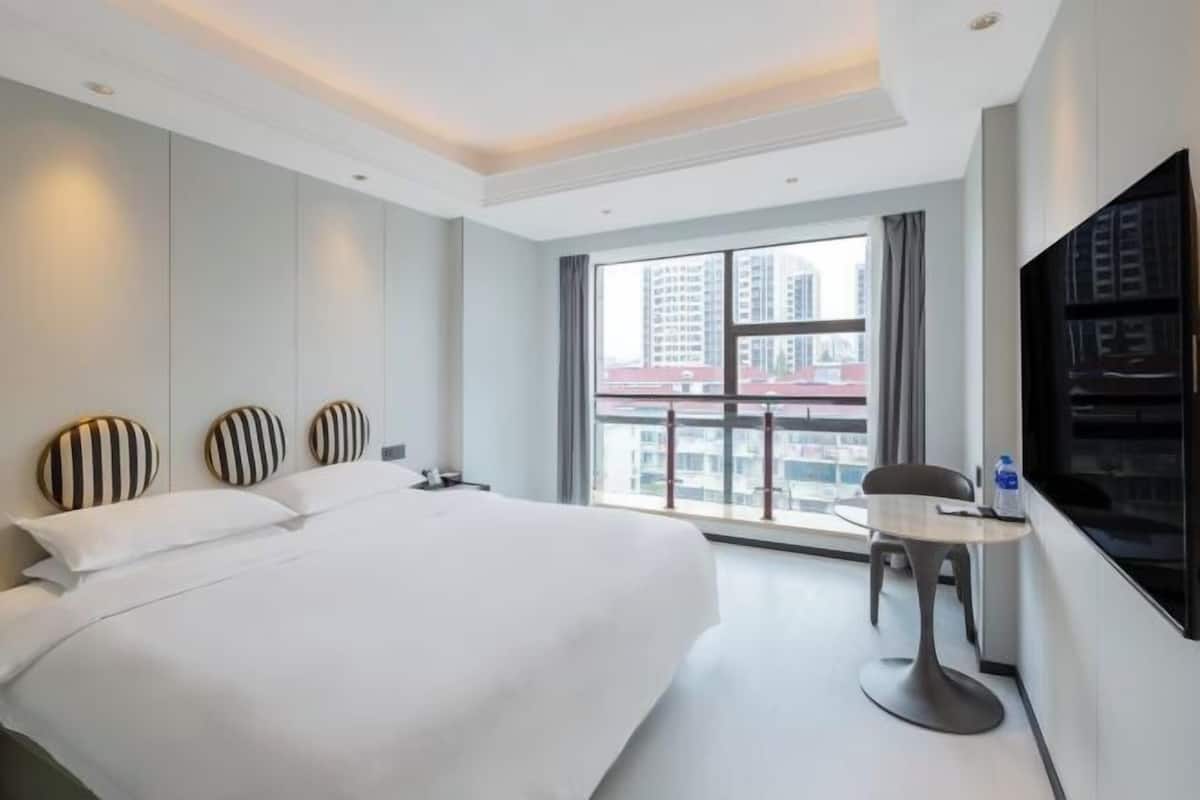 A hotel room with a large bed, a small table, a chair, a TV, and a window with a city view.