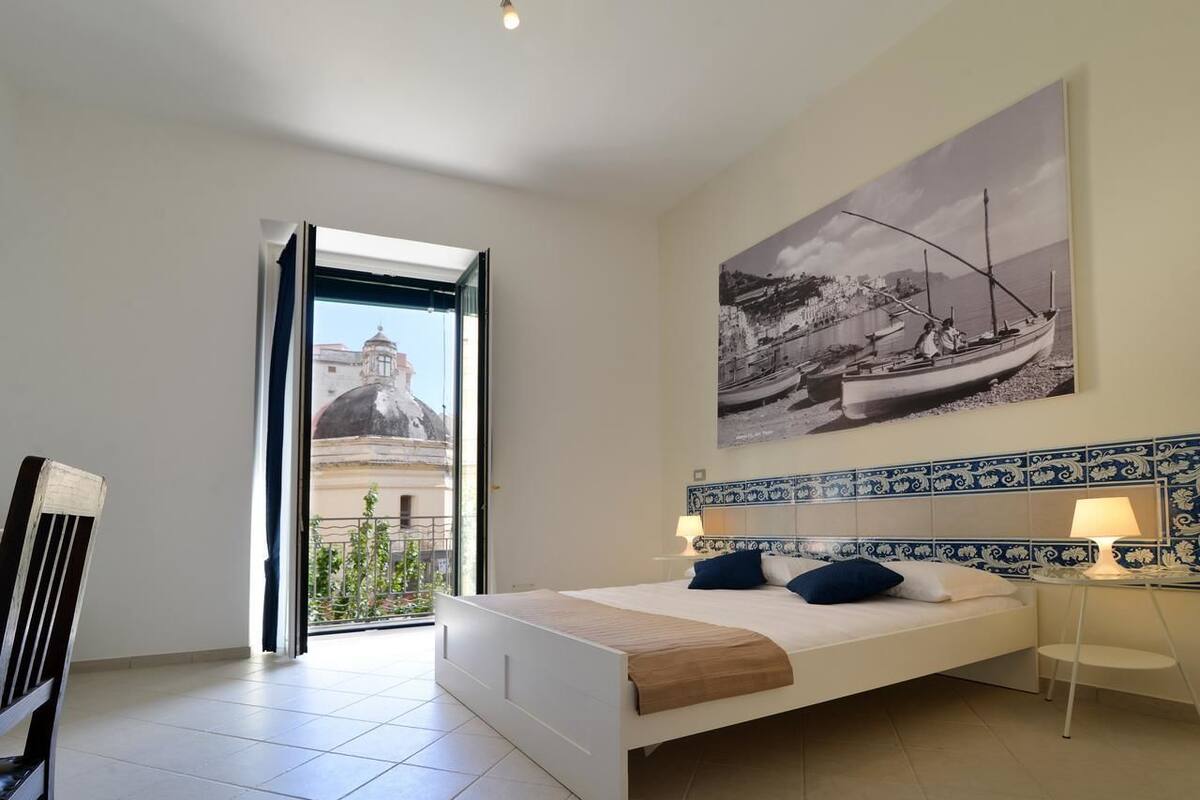 A bedroom with a bed, a nightstand, a lamp, and a large window with a view of a building and a boat.