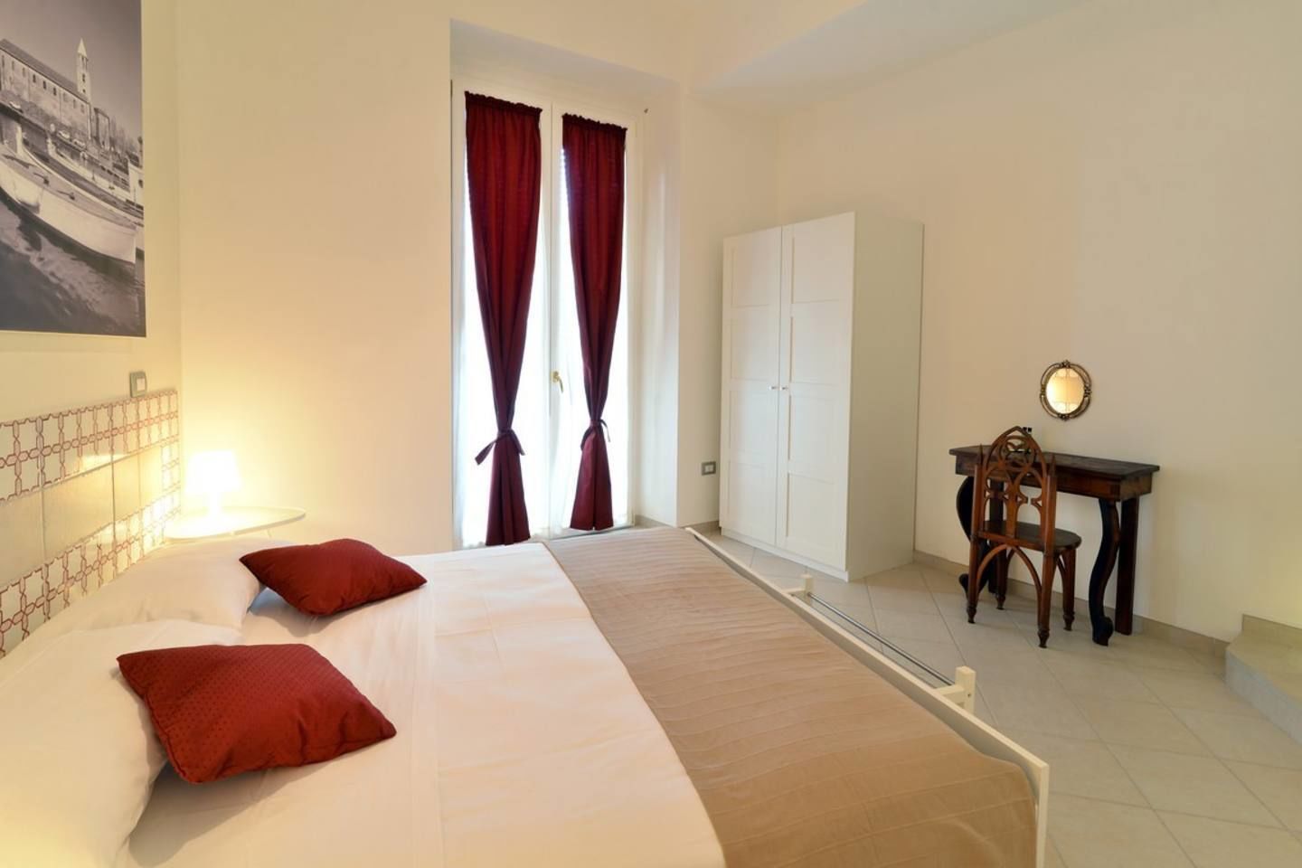 A hotel room with a bed, two red pillows, a desk with a chair, a wardrobe, and a window with curtains.