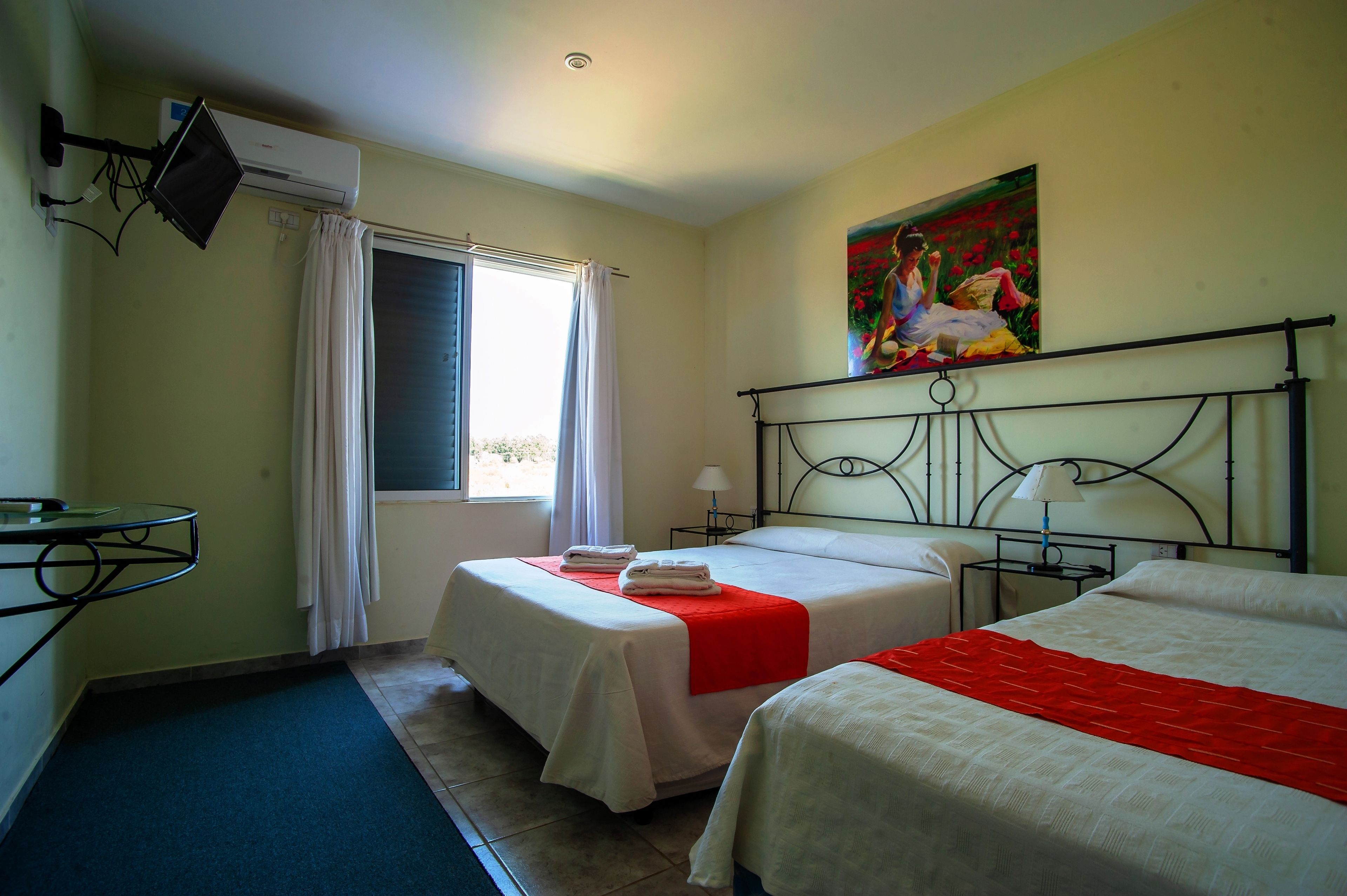A hotel room with two beds, a window with curtains, an air conditioning unit, a painting on the wall, and a small table with a lamp.