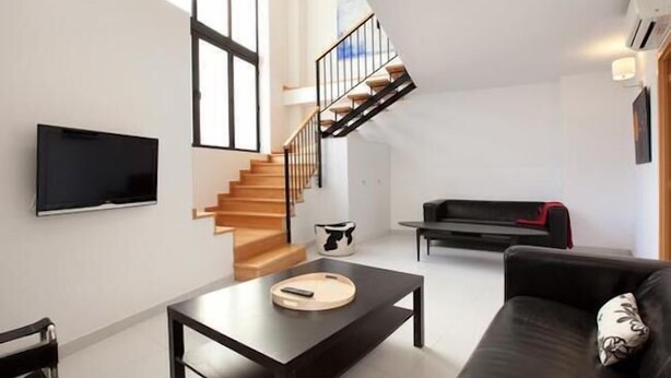 A modern living room with a staircase, a flat-screen TV mounted on the wall, a black sofa, and a coffee table. - Park Guell Apartments (Barcelona)