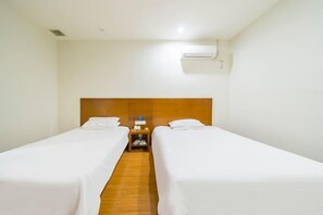 A hotel room with two single beds, a wooden headboard, a nightstand with a lamp, and an air conditioning unit.
