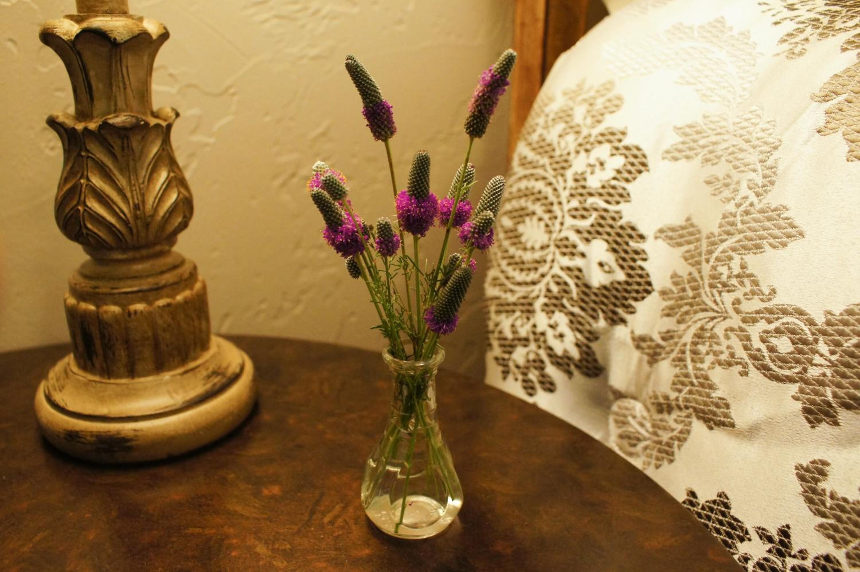 Um vaso de vidro transparente com flores roxas sobre uma mesa de madeira, ao lado de um abajur decorativo e uma almofada estampada.