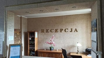 Zona de recepción con mostrador, un cartel en la pared que dice 'RECEPCJA', una orquídea en maceta, un calendario y una estantería con bebidas embotelladas.