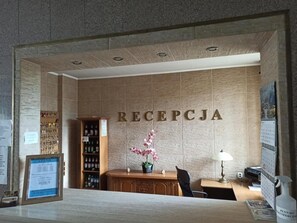 A reception area with a reception desk, a wall-mounted sign reading 'RECEPCJA', a potted orchid, a calendar, and a shelf with bottled beverages.