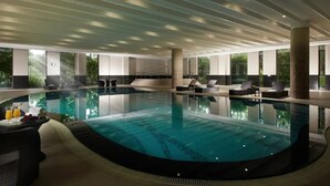 Ein moderner, innenliegender Schwimmbereich mit geschwungener Form, umgeben von Liegebereichen, Sitzgelegenheiten und einem Tisch mit Getränken und Früchten.