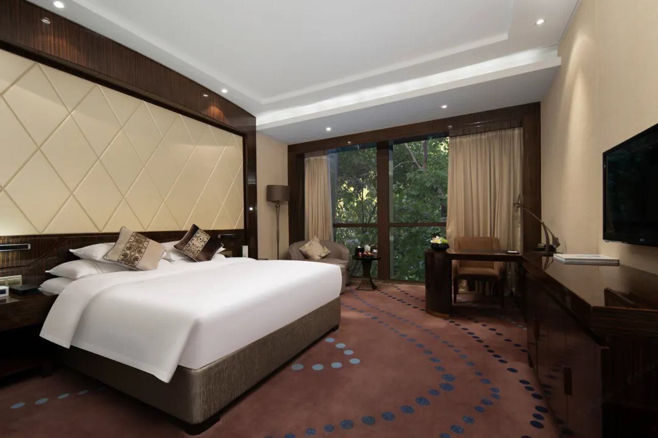 A hotel room with a large bed, a desk, a chair, a TV, and a balcony with curtains.