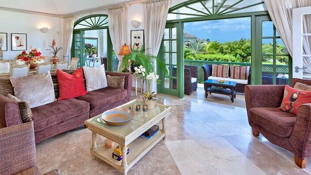 A spacious living room with a sofa, armchairs, and a coffee table. There is a dining area with a table and chairs. The room has large windows with a view of greenery outside. - The Summer House, Sugar Hill Resort (Holetown)
