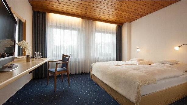 Quarto de hotel com teto de madeira, cama com roupa de cama branca, escrivaninha com cadeira e uma janela grande com cortinas.
