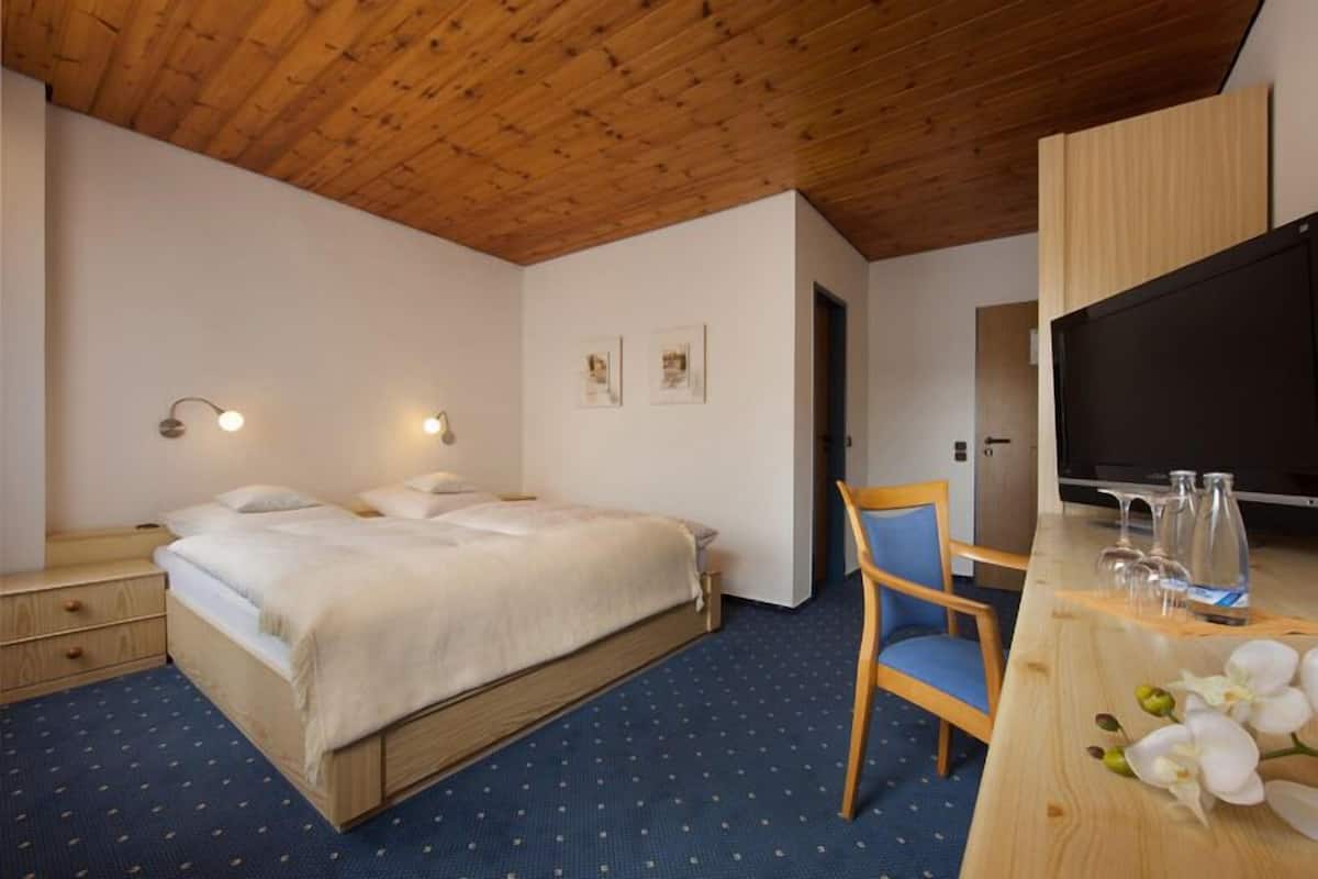 A hotel room with a wooden ceiling, a bed with white bedding, a desk with a chair, and a television.