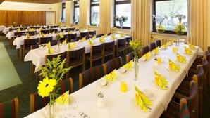 Uma sala de banquetes com várias mesas redondas preparadas para refeições, com guardanapos amarelos e centros de mesa florais.