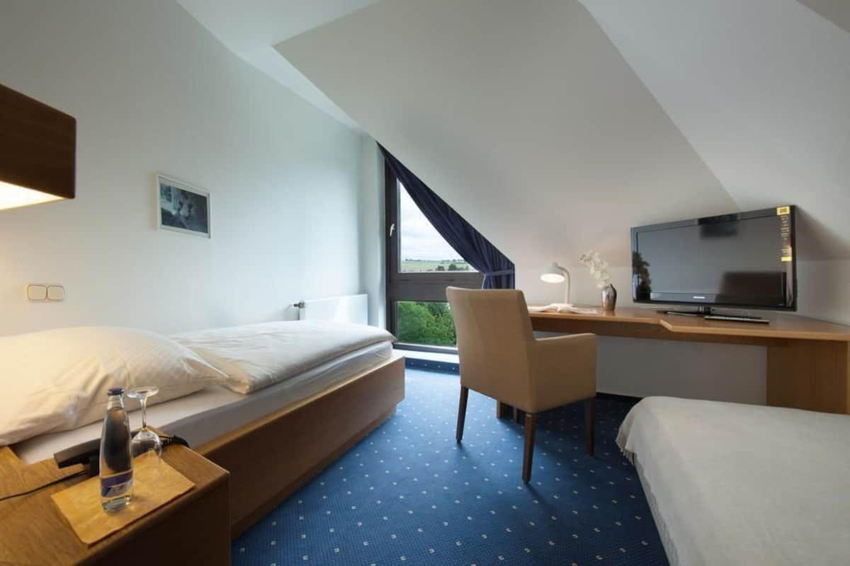 A hotel room with a bed, a desk with a TV, a chair, and a window with curtains.