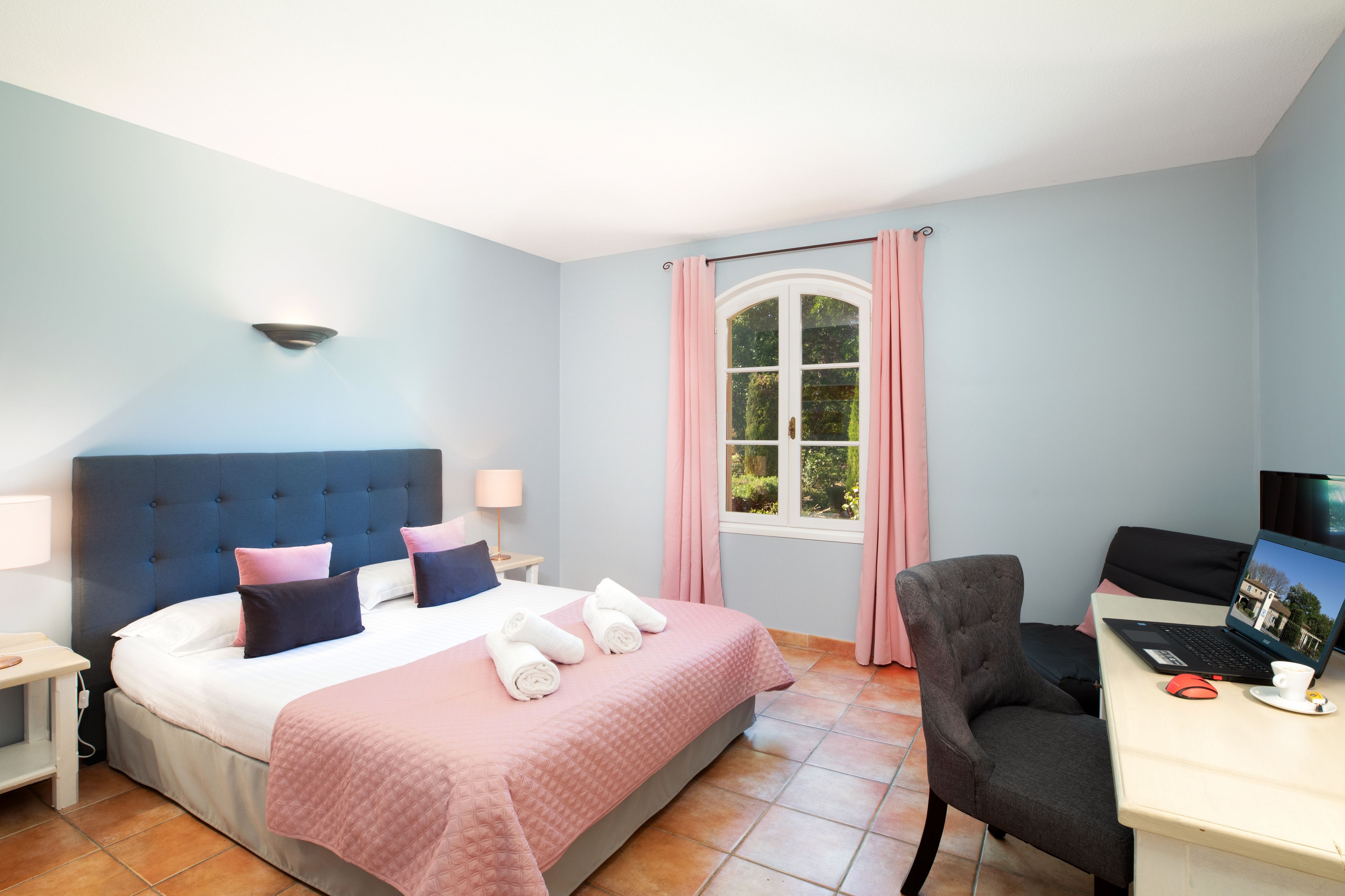 A bedroom with a bed, a desk with a laptop, a chair, and a window with curtains.