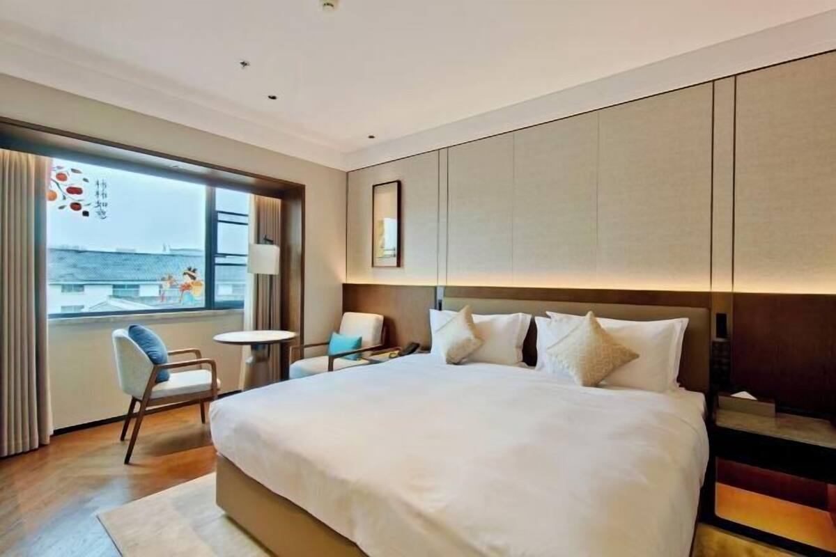 A modern hotel room with a large bed, a small table, two chairs, a view of the sea, and a large window.