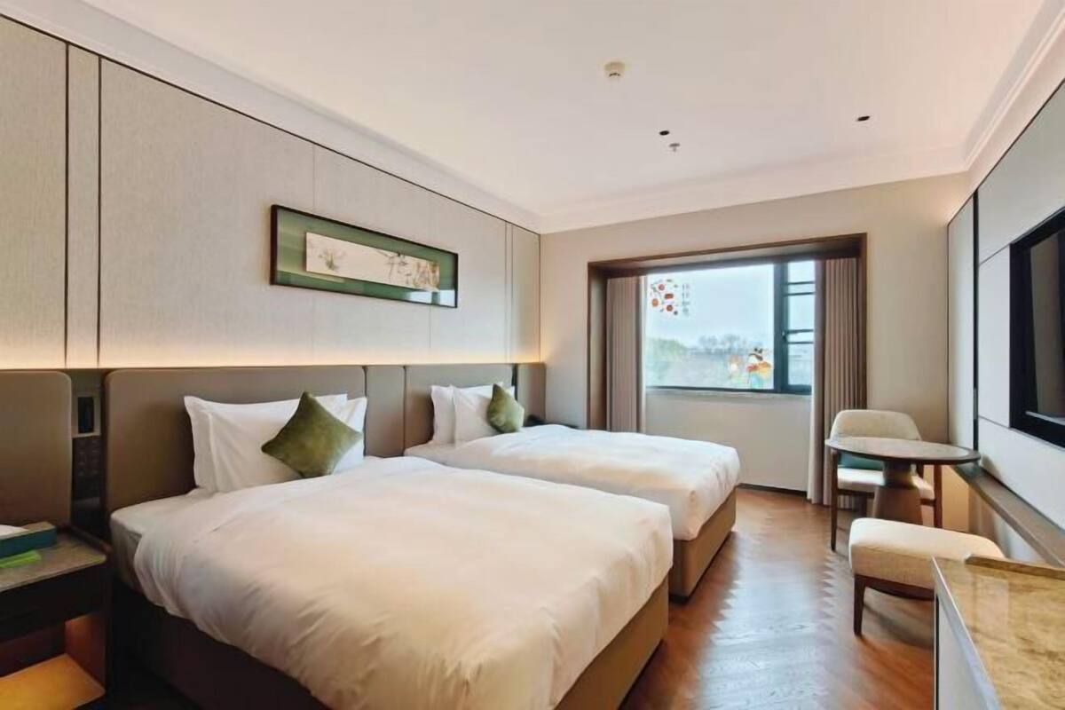 A hotel room with two beds, a small table, a chair, and a window with a view.