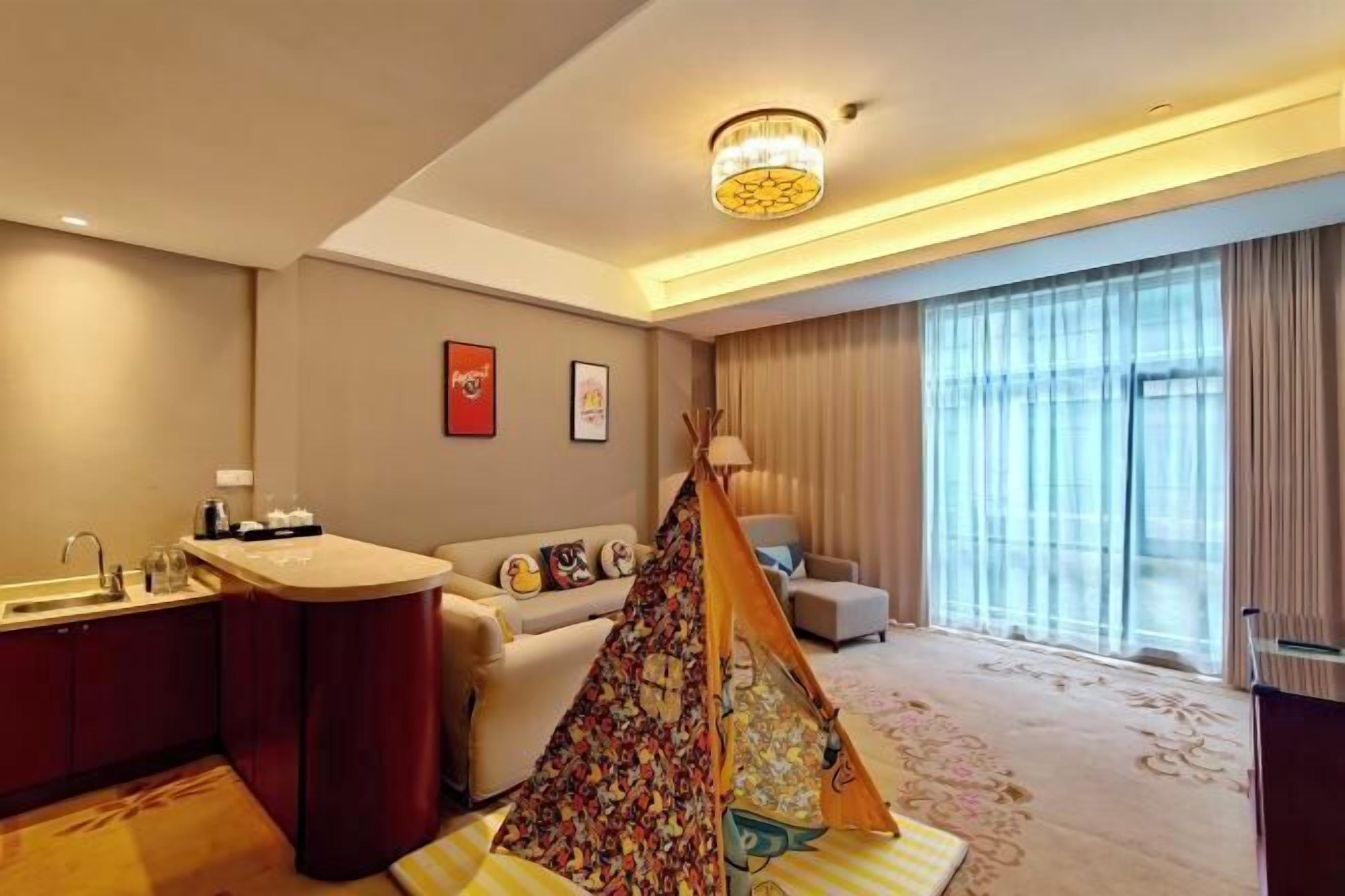 Une chambre d’hôtel équipée d’un coin cuisine, d’un canapé, d’un espace de jeu aménagé comme une tente, et d’une grande fenêtre avec des rideaux.