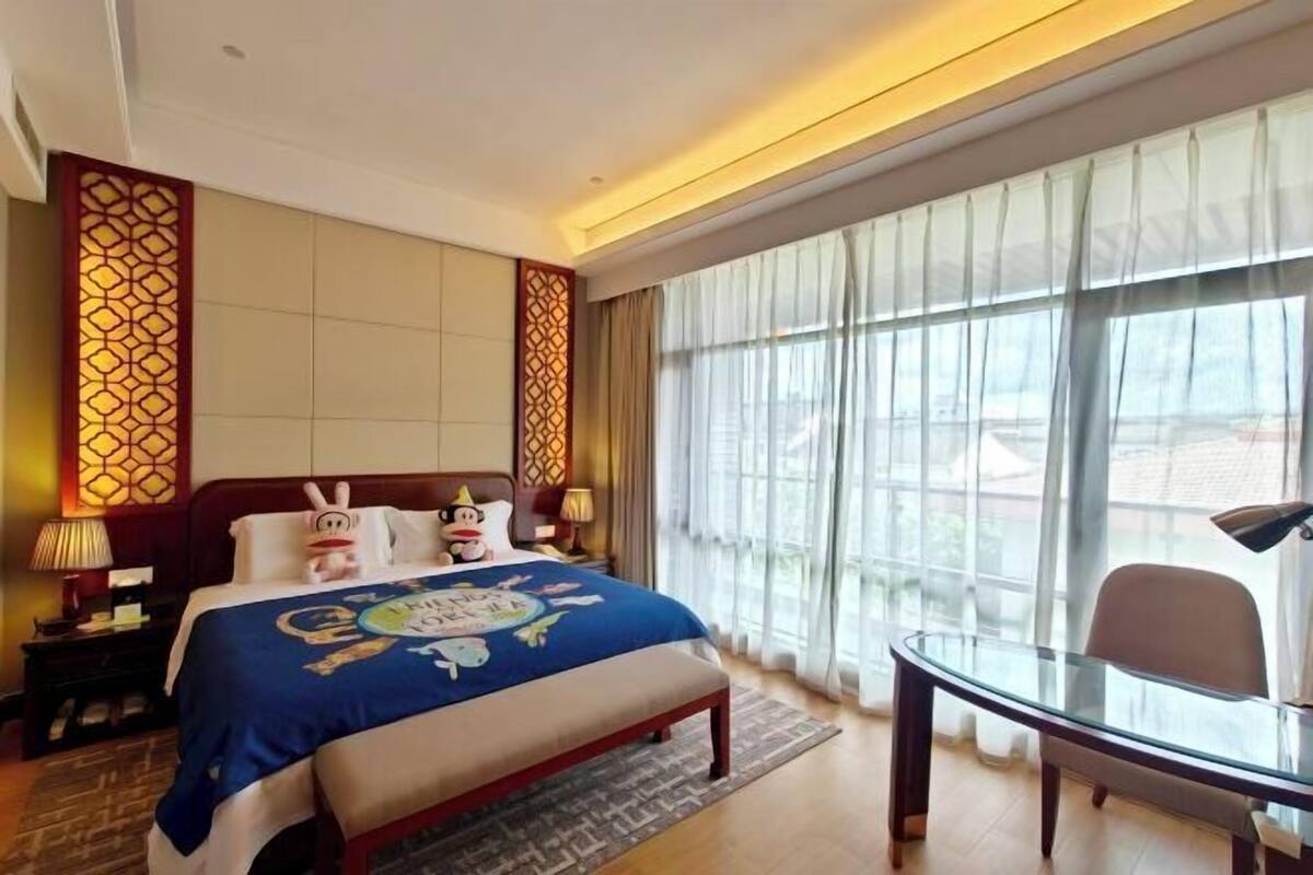 A hotel room with a bed, bedside tables, a desk, and large windows.