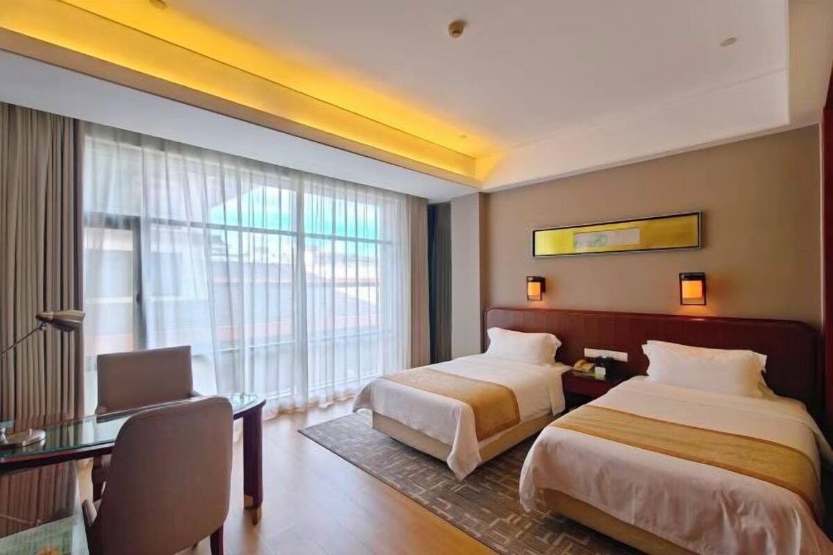 A hotel room with two beds, a desk, a chair, a lamp, and a large window with sheer curtains.