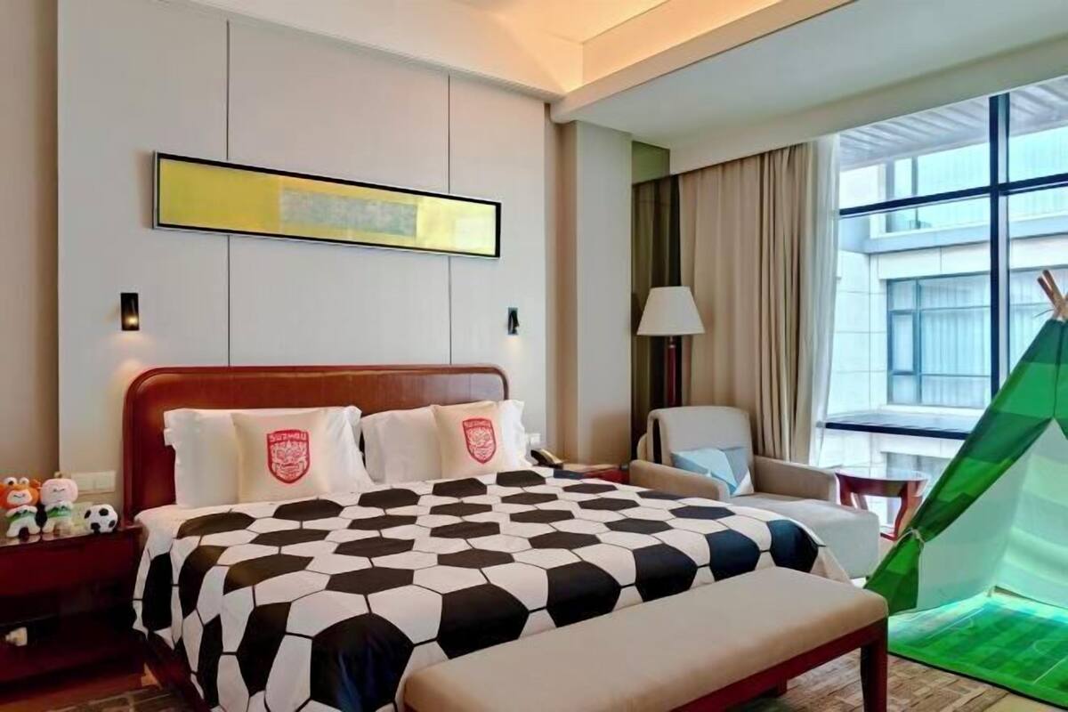 A hotel room with a large bed, a chair, a lamp, a green tent, and a window with a view of buildings.