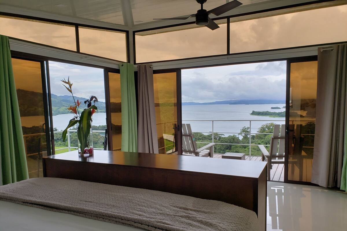 A bedroom with a large bed, a view of a lake, and a balcony with chairs.