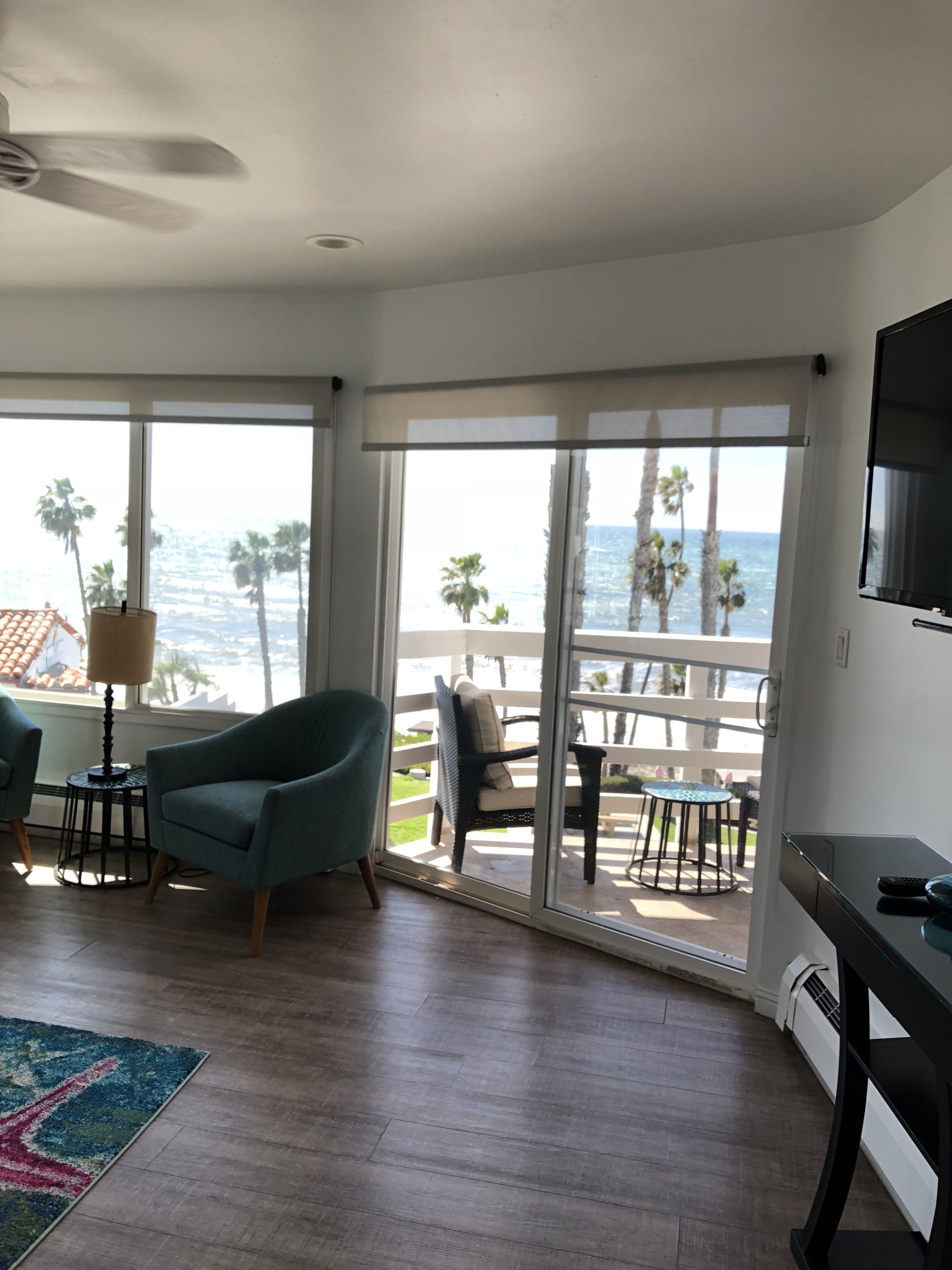 A room with a large sliding glass door leading to a balcony with a view of the ocean and palm trees. There are two chairs and a small table with a lamp.