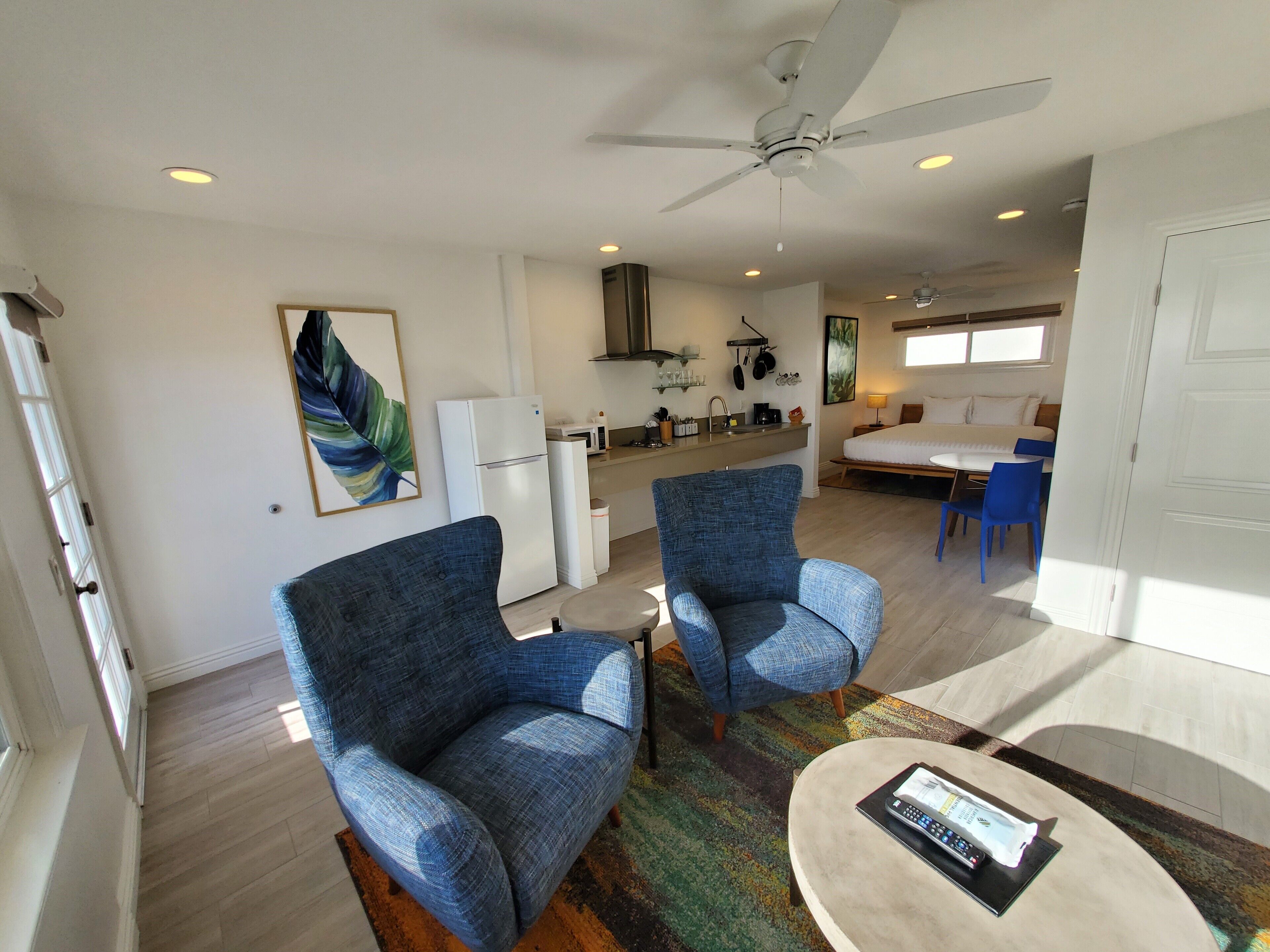 A modern living room with two blue armchairs, a coffee table, a ceiling fan, and a kitchen area in the background.