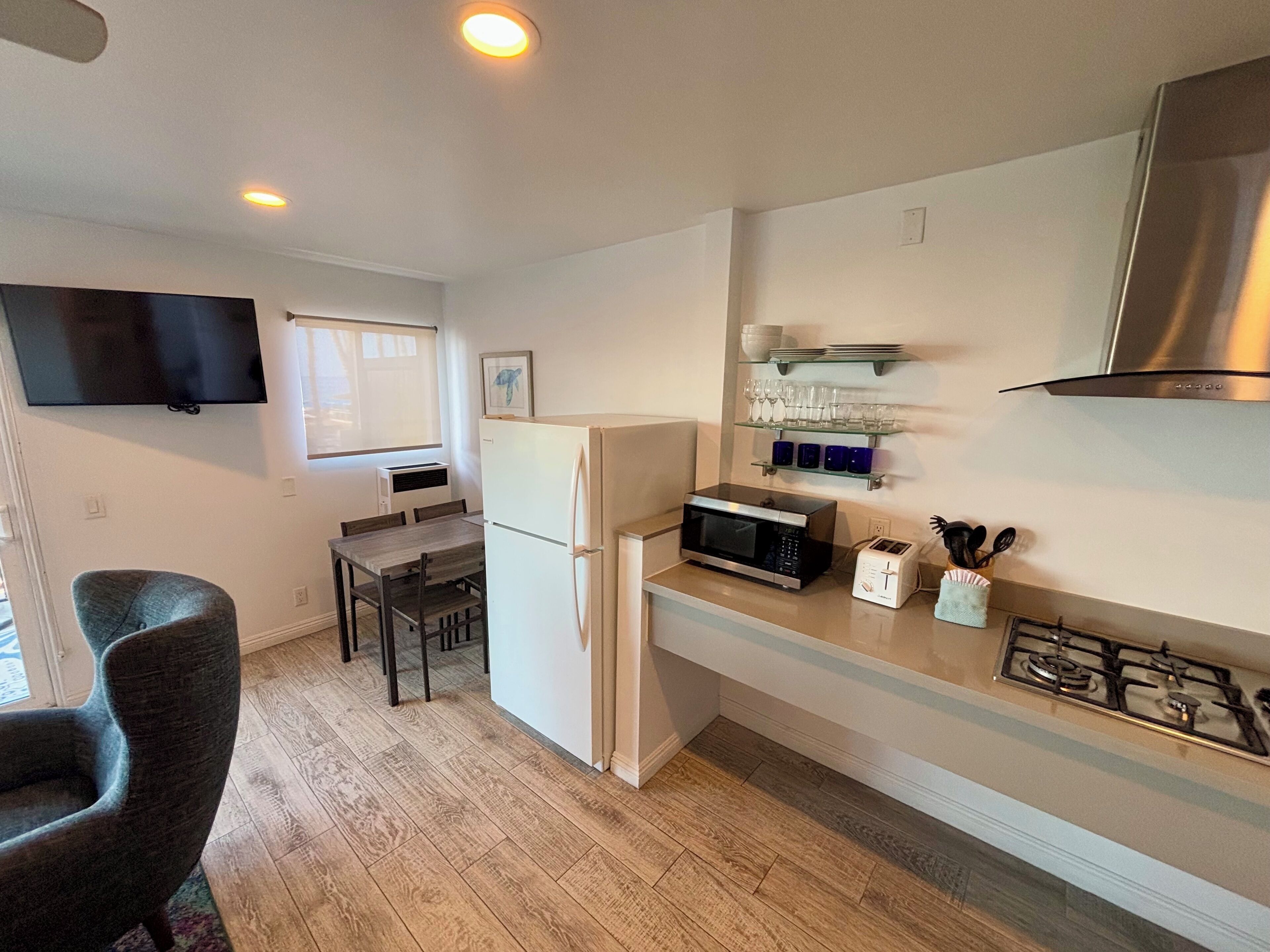 A compact kitchen with a refrigerator, microwave, and gas stove. There is a dining table with chairs and a flat-screen TV mounted on the wall.