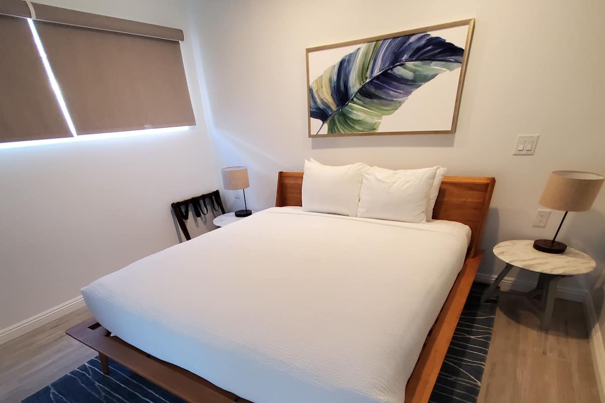 Ein modernes Schlafzimmer mit einem großen Bett, einem dunklen Teppich mit Muster, einem Holz-Kopfteil, einem an der Wand hängenden Kunstwerk und einem kleinen Nachttisch mit Lampe.